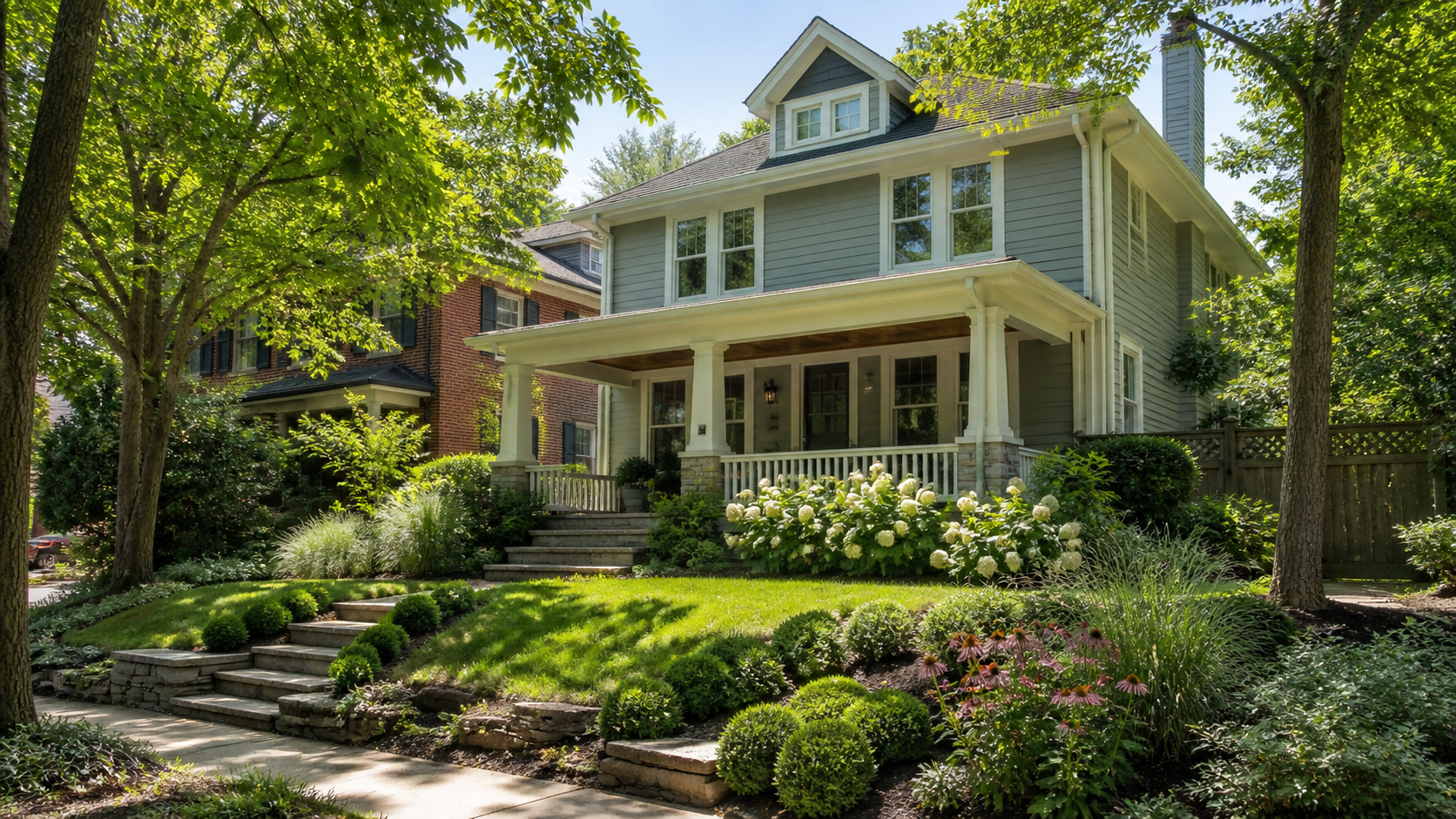 Two-story blue-gray house with a covered front porch, white columns, and stone accents, surrounded by lush landscaping and white hydrangeas in bloom.